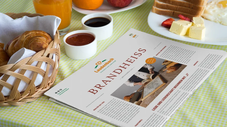 Kundenmagazin „Brandheiss“ der Bäckerei Laudenbach auf einem Frühstückstisch mit Croissants, Butter und Kaffee.