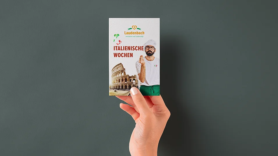 Hand hält Hangtag der Bäckerei Laudenbach für die Italienischen Wochen mit italienischem Bäcker und Kolosseum-Motiv.