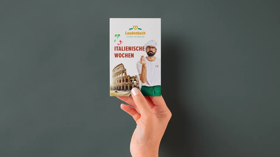 Hand hält Hangtag der Bäckerei Laudenbach für die Italienischen Wochen mit italienischem Bäcker und Kolosseum-Motiv.