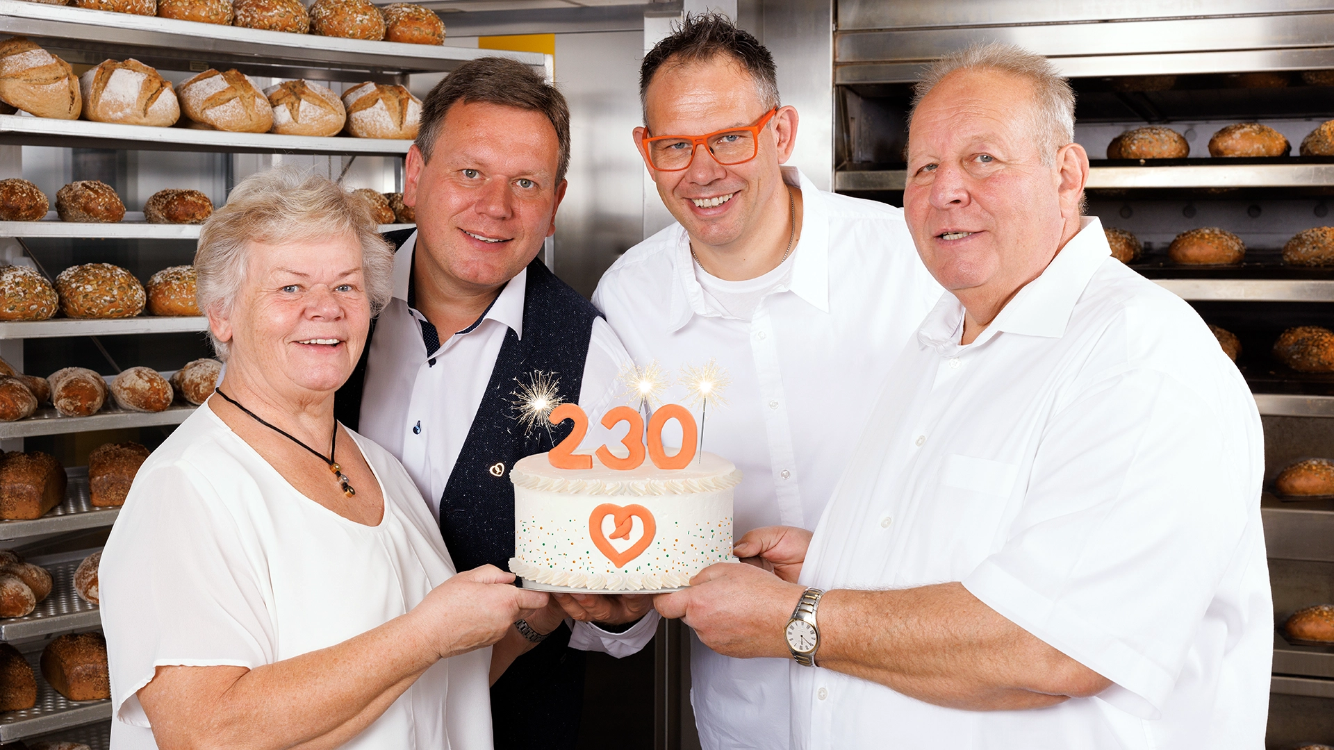 Vier Personen halten gemeinsam eine Jubiläumstorte mit der Zahl 230 und Herz-Logo, im Hintergrund frisches Brot in der Backstube